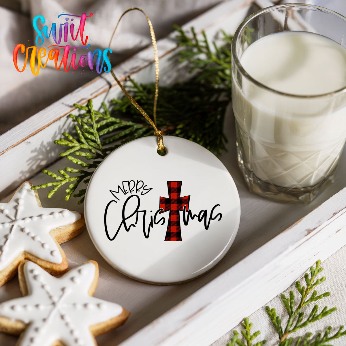 a festive Christmas scene with a white ceramic ornament hanging from a string, displaying the text "Merry Christmas" in a stylized font. The ornament is surrounded by a glass of milk, cookies, and greenery, creating a cozy and holiday atmosphere.