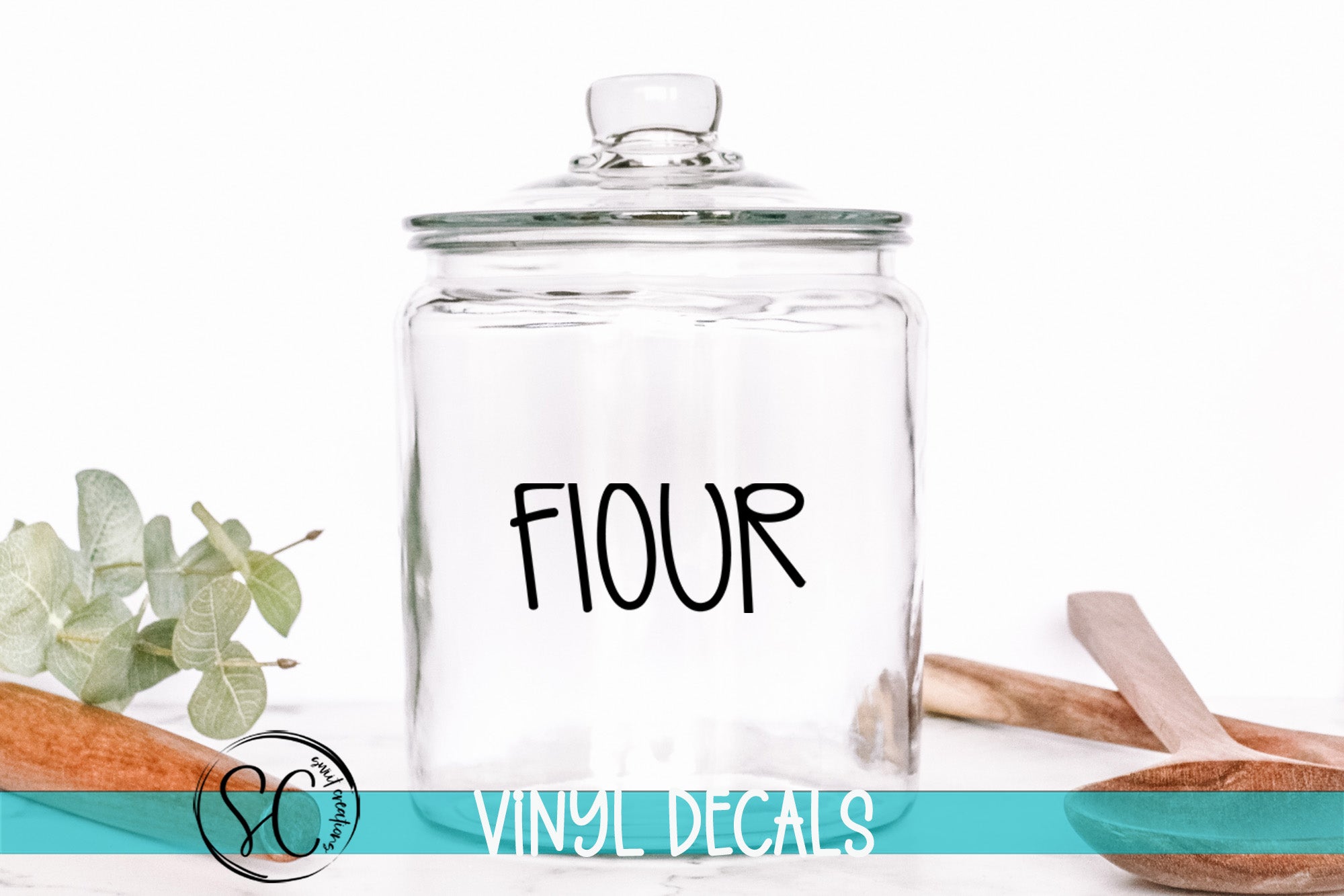 A clear glass jar with the word "FLOUR" printed on it, placed on a white surface next to some wooden utensils.