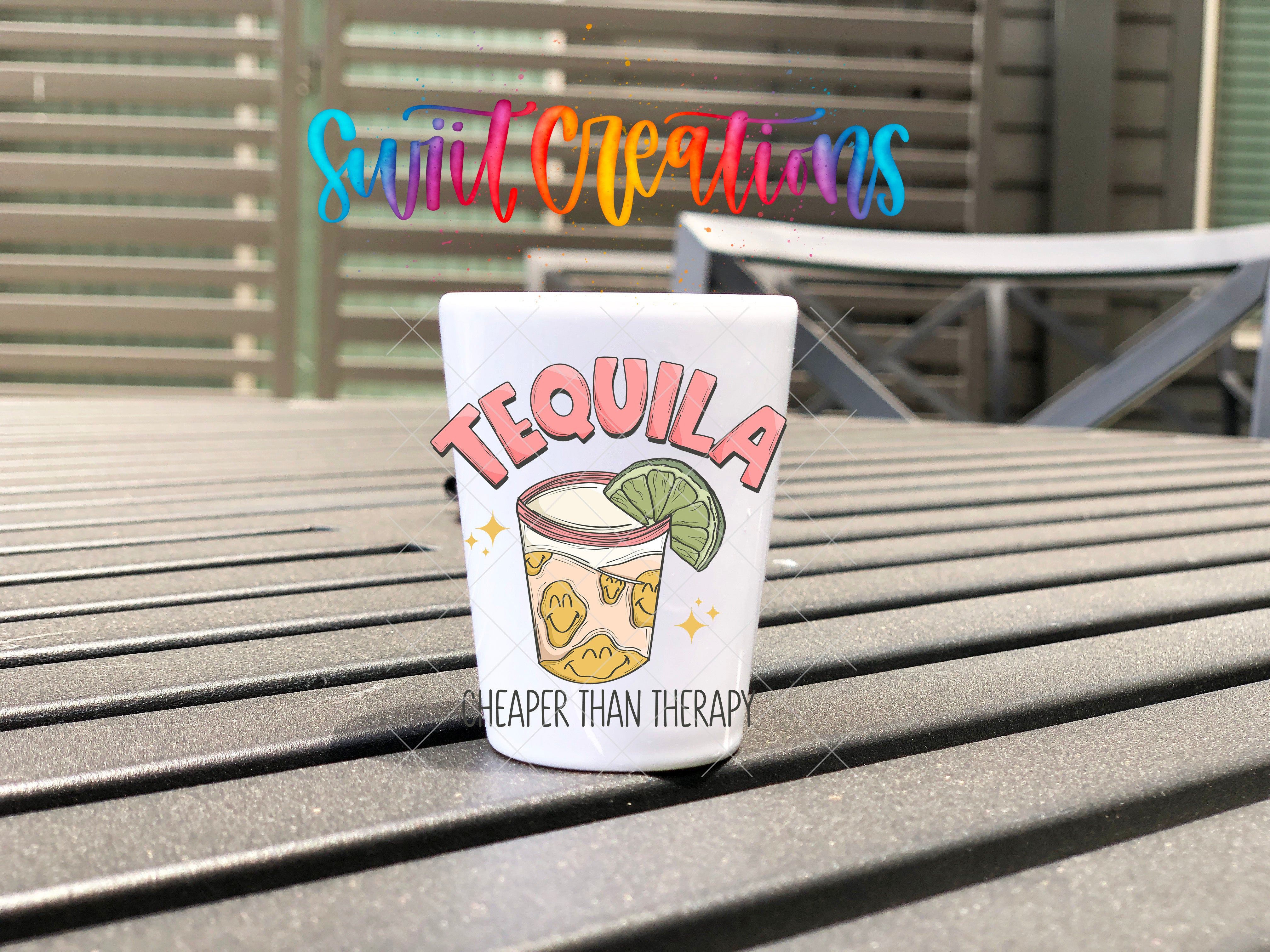 A white tequila shot mug with a colorful design and text, placed on a wooden surface.