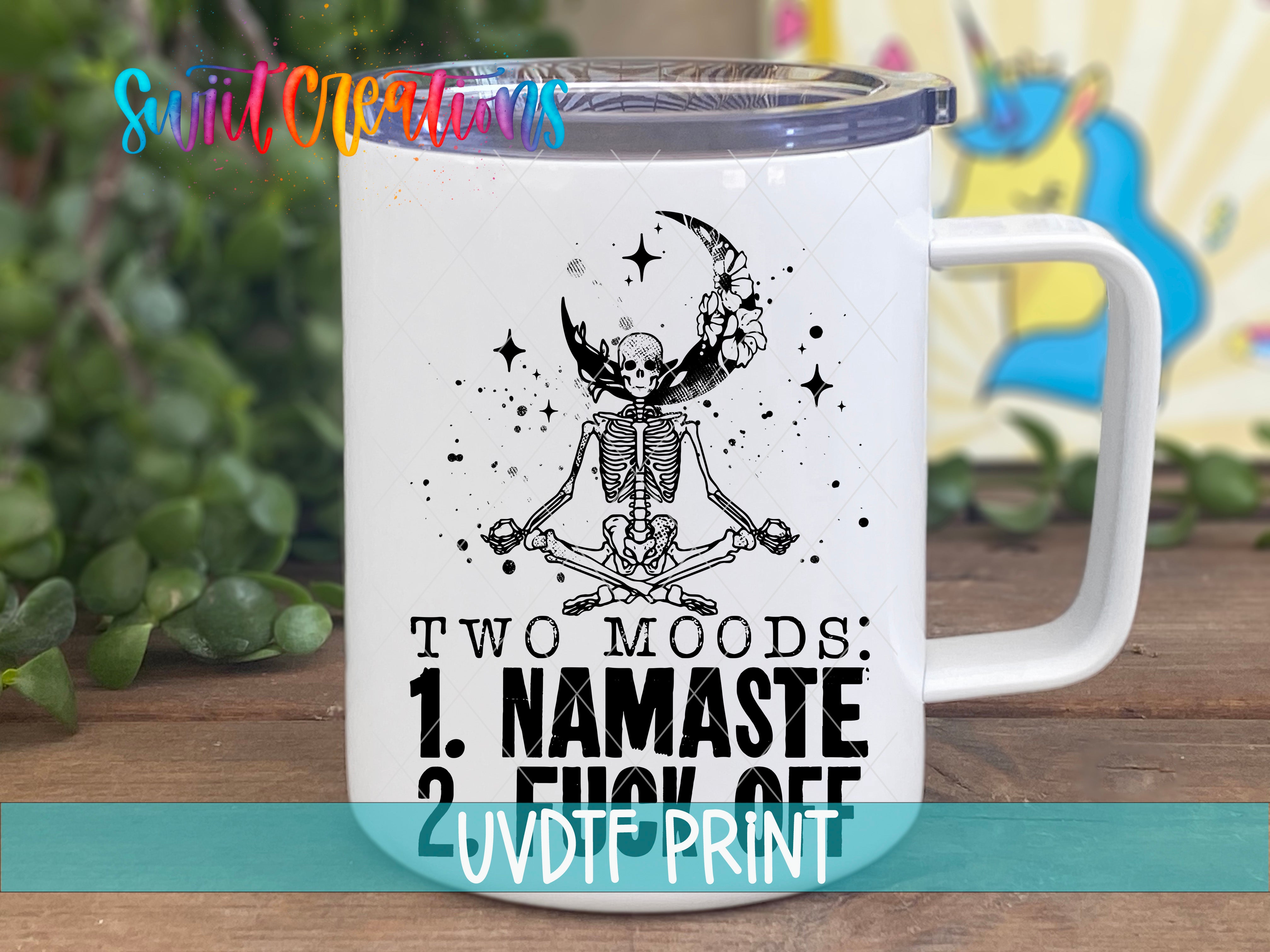 A white mug with a black and white illustration of a skeleton in a meditative pose, surrounded by stars and a crescent moon. The mug has text that reads "Two moods: 1. NAMASTE 2. FICK OFF" and "UVDIF PRINT".