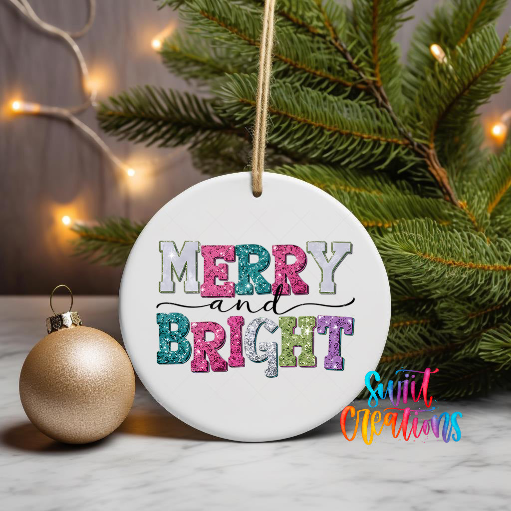 A white ceramic ornament with the text "MERRY AND BRIGHT" in colorful letters, hanging from a string. The ornament is placed next to a gold ornament and a pine tree, with a string of lights visible in the background.