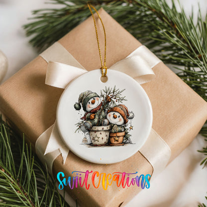 A ceramic ornament featuring two snowmen sitting in a pot, surrounded by pine branches, is displayed on a gift box with a white ribbon.