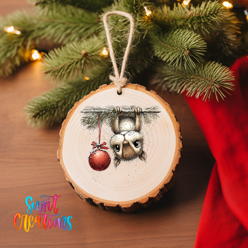 A wooden ornament with a picture of a cat hanging from a branch, holding a red ornament, is displayed on a Christmas tree.