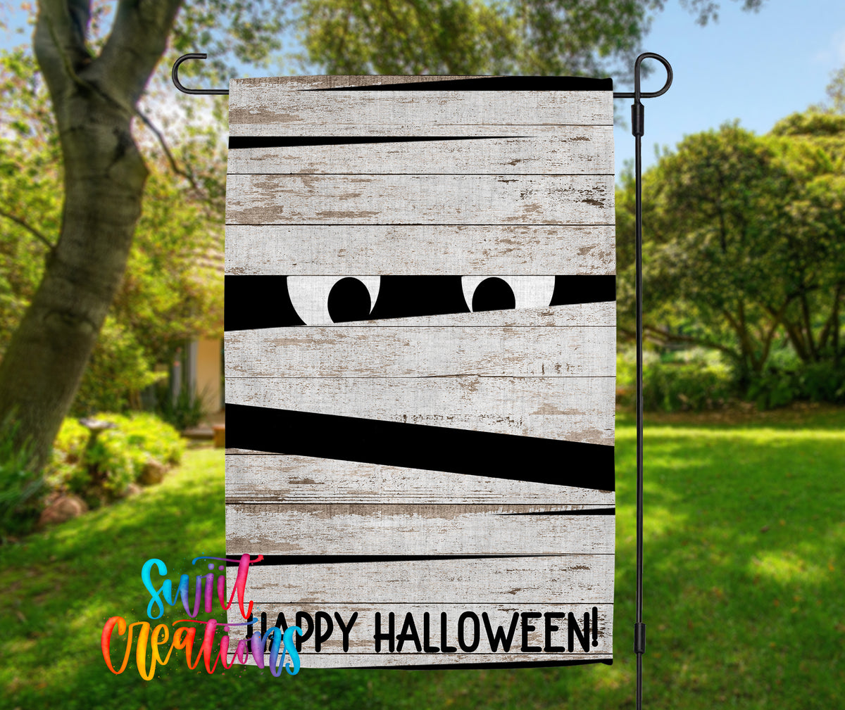 A Halloween-themed garden decoration with a wooden board displaying a mummified face and the text "Happy Halloween!"