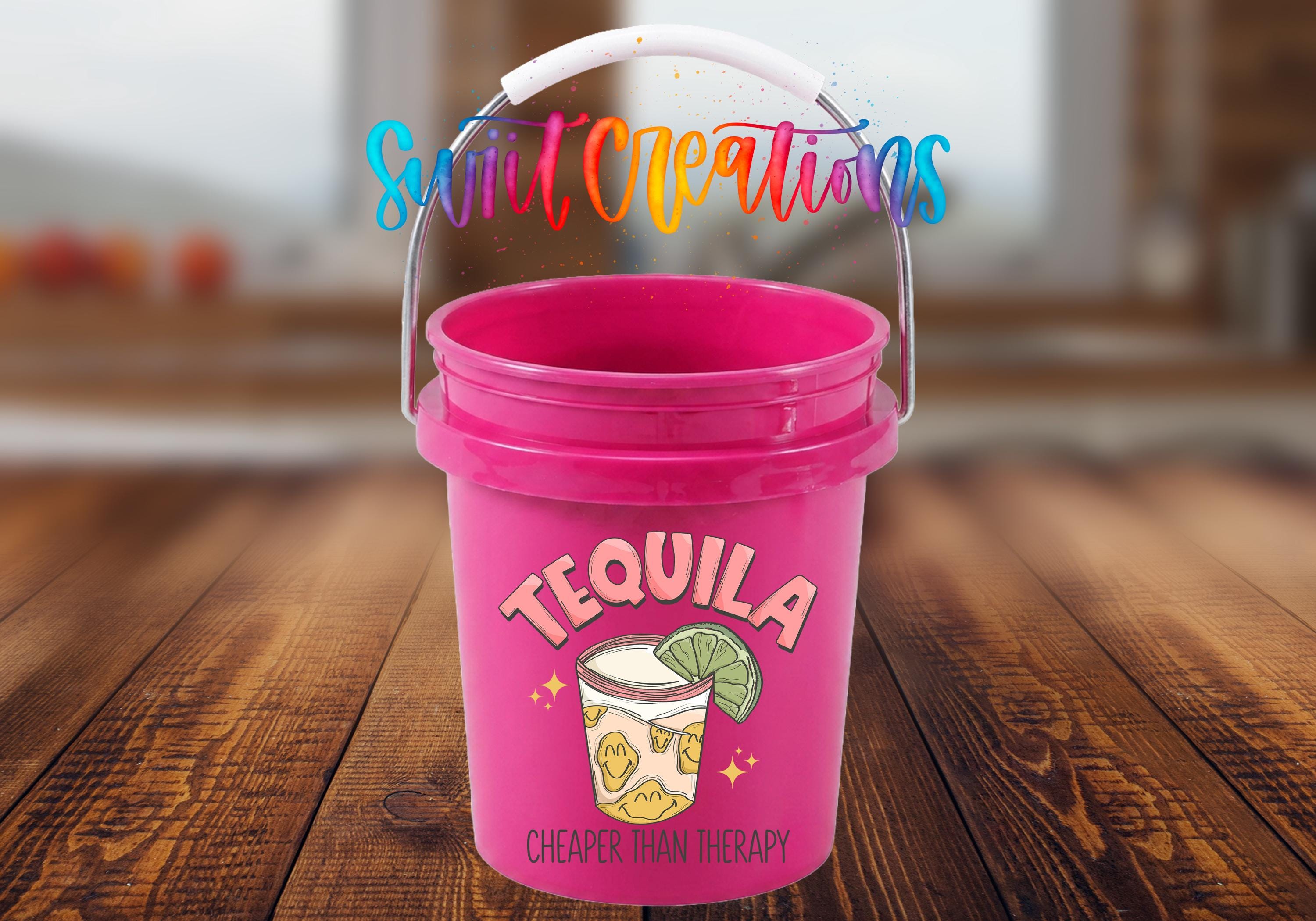 A pink bucket with a tequila-themed design and the text "Tequila" and "CHEAPER THAN THERAPY" printed on it, placed on a wooden surface.