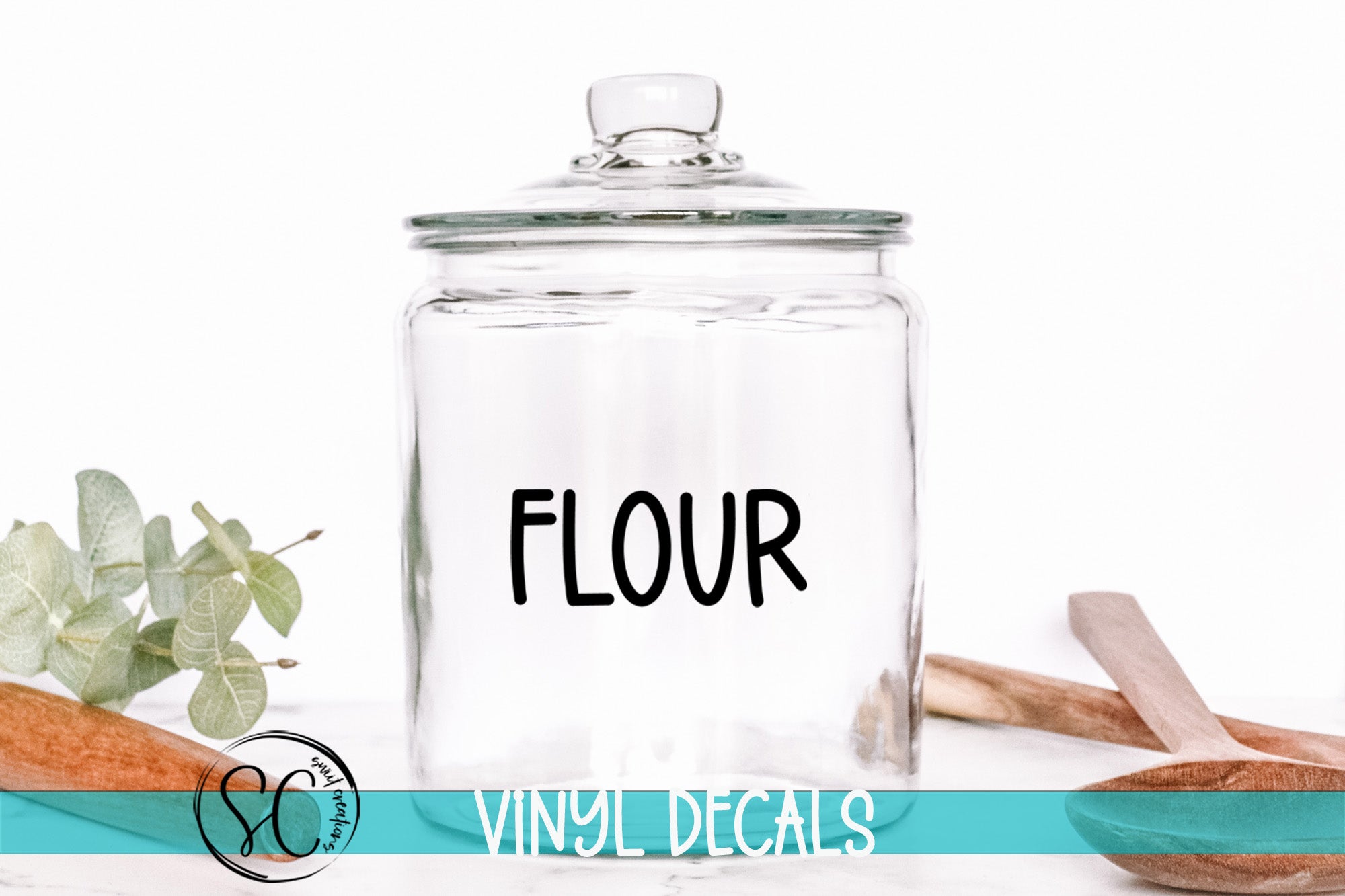 A clear glass jar with the word "FLOUR" printed on it, placed on a white surface next to some wooden utensils.
