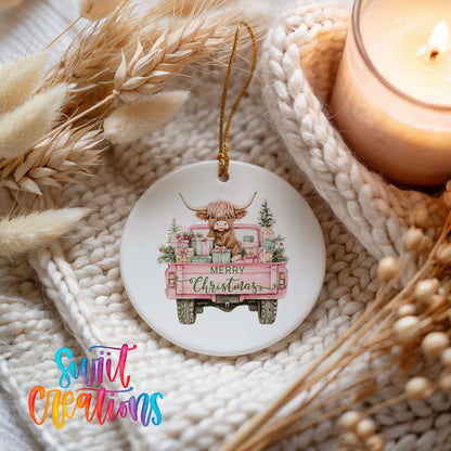 A ceramic ornament featuring a cartoon cow in a pink truck, with the text "Merry Christmas" displayed on the truck. The ornament is placed on a white knitted surface, with dried flowers and a lit candle nearby.