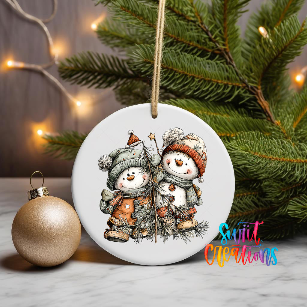 A white ceramic ornament featuring two snowmen, one wearing a red hat and the other a green hat, is displayed on a table next to a gold ornament and a Christmas tree.
