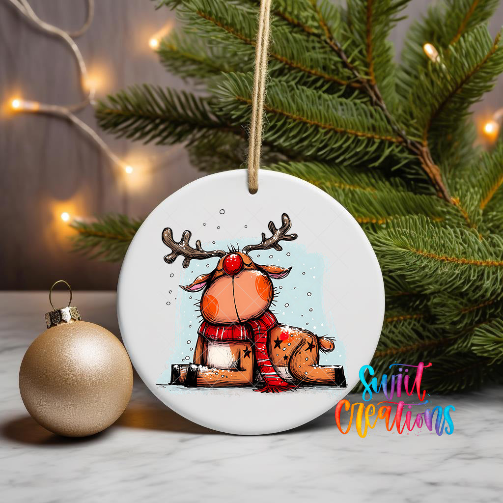 A white ceramic ornament featuring a cartoon reindeer with antlers and a red scarf, sitting on a snowy surface, is displayed in front of a Christmas tree with twinkling lights.