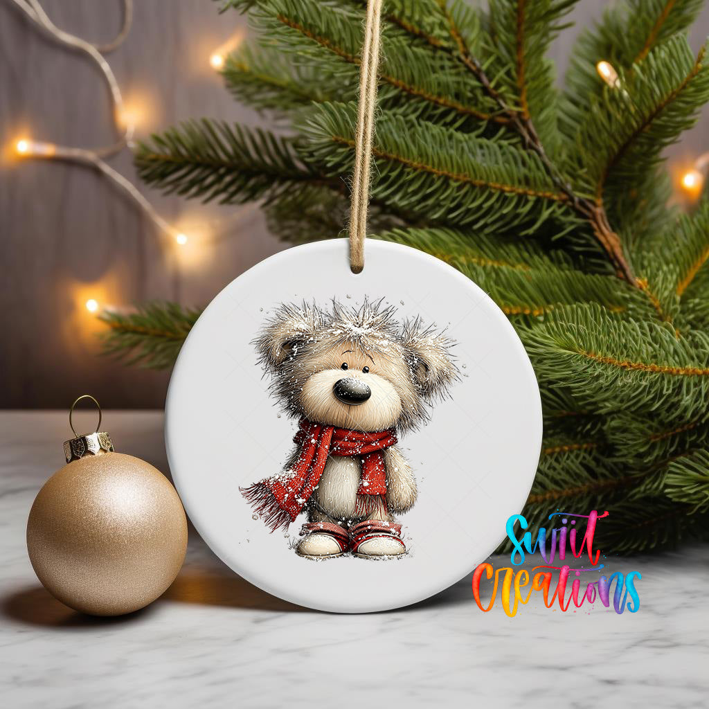 A white ceramic ornament featuring a teddy bear wearing a red scarf, hanging on a string next to a gold ornament and a Christmas tree.