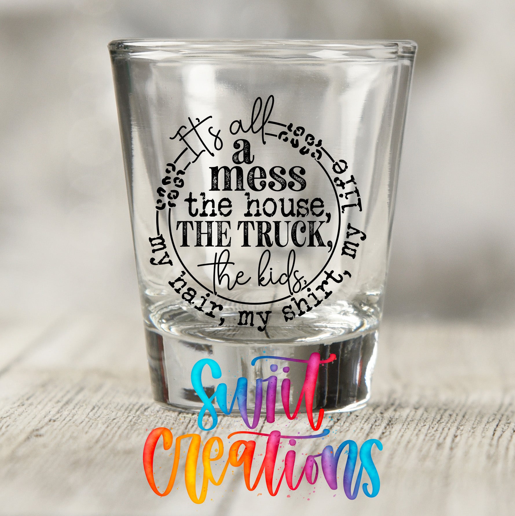 A shot glass with a humorous text printed on it, sitting on a wooden surface.