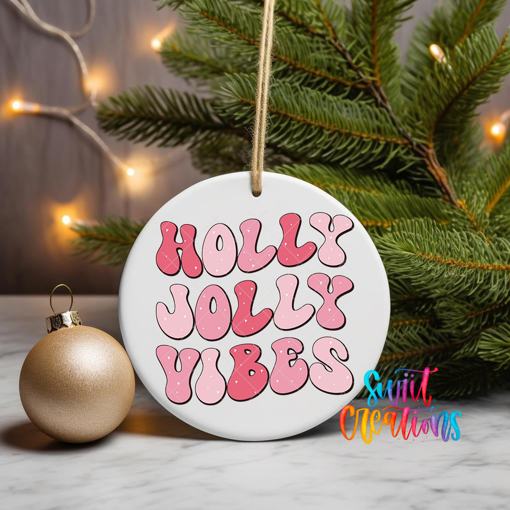 A white ceramic ornament with the text "HOLY JOYLY VIBES" in pink and red letters, hanging from a string. The ornament is placed next to a gold ornament on a marble surface, with a pine tree and twinkling lights in the background.