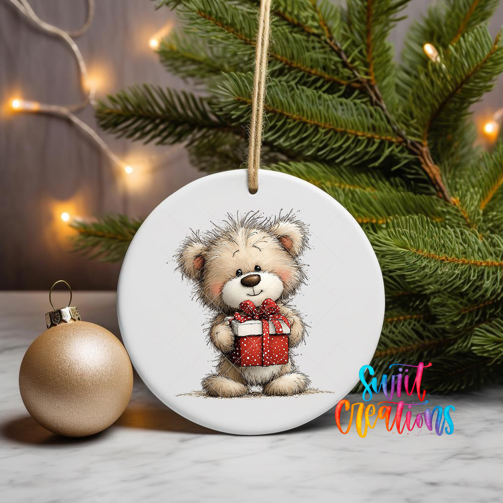 A white ceramic ornament featuring a teddy bear holding a red gift box, placed on a marble surface next to a gold ornament and a Christmas tree with twinkling lights.