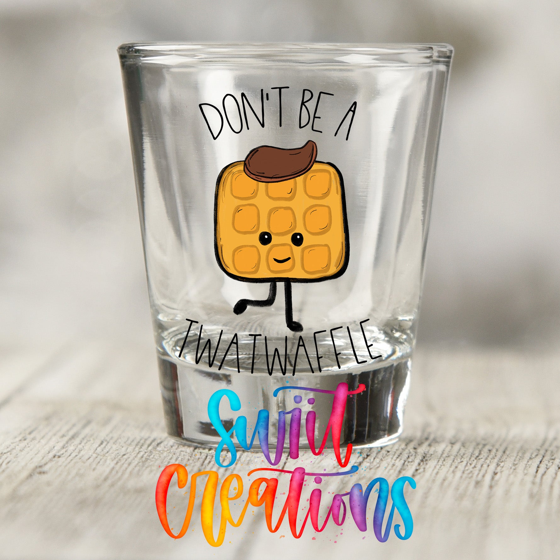 a shot glass with a cartoon waffle on it, along with the text "DON'T BE A TWATWACKEL" and "SWEET CREATIONS" written below it.