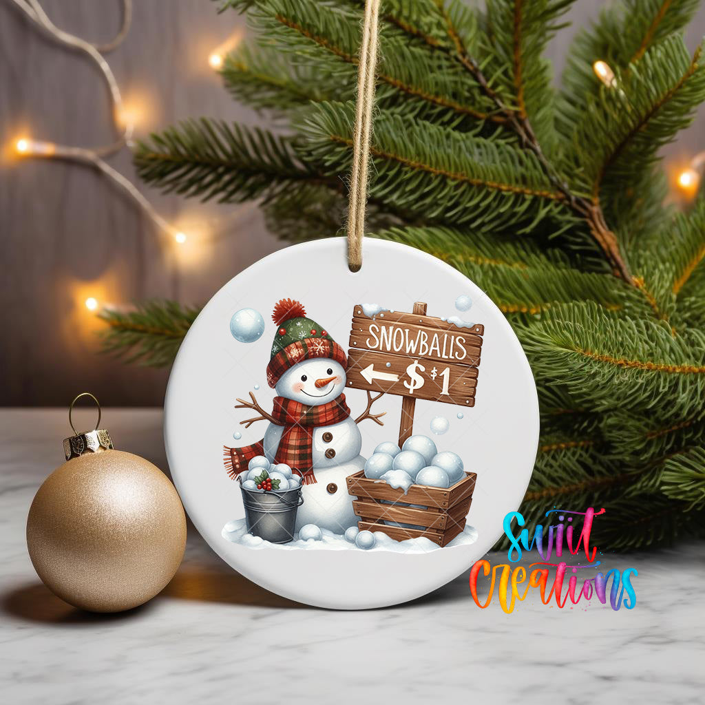 A snowman ornament with a sign that says "Snowballs $1" is displayed on a Christmas tree, with a gold ornament and a pine tree in the background.