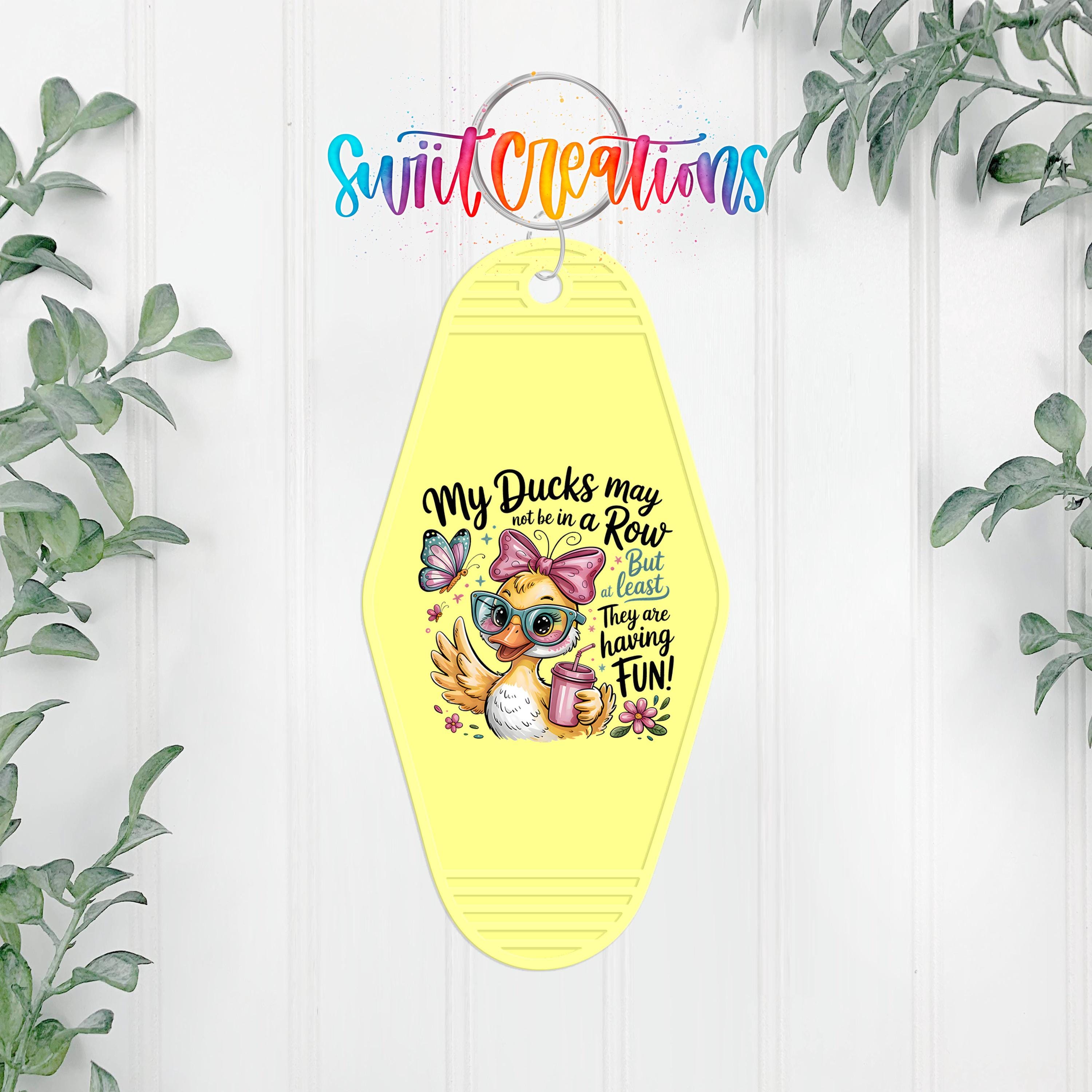 A yellow keychain with a cartoon duck and text, hanging on a white wall with greenery in the background.