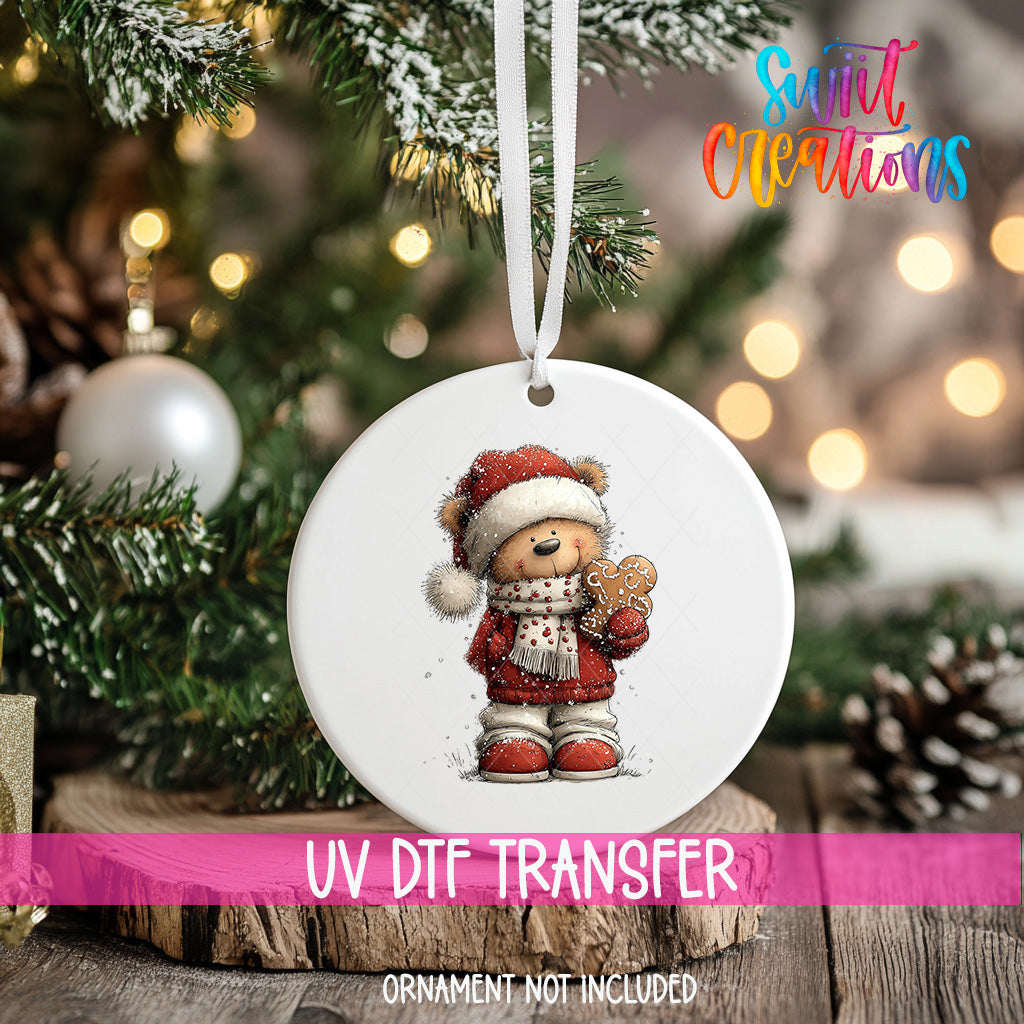 A white ornament with a teddy bear wearing a Santa hat and holding a gingerbread man is hanging on a Christmas tree.