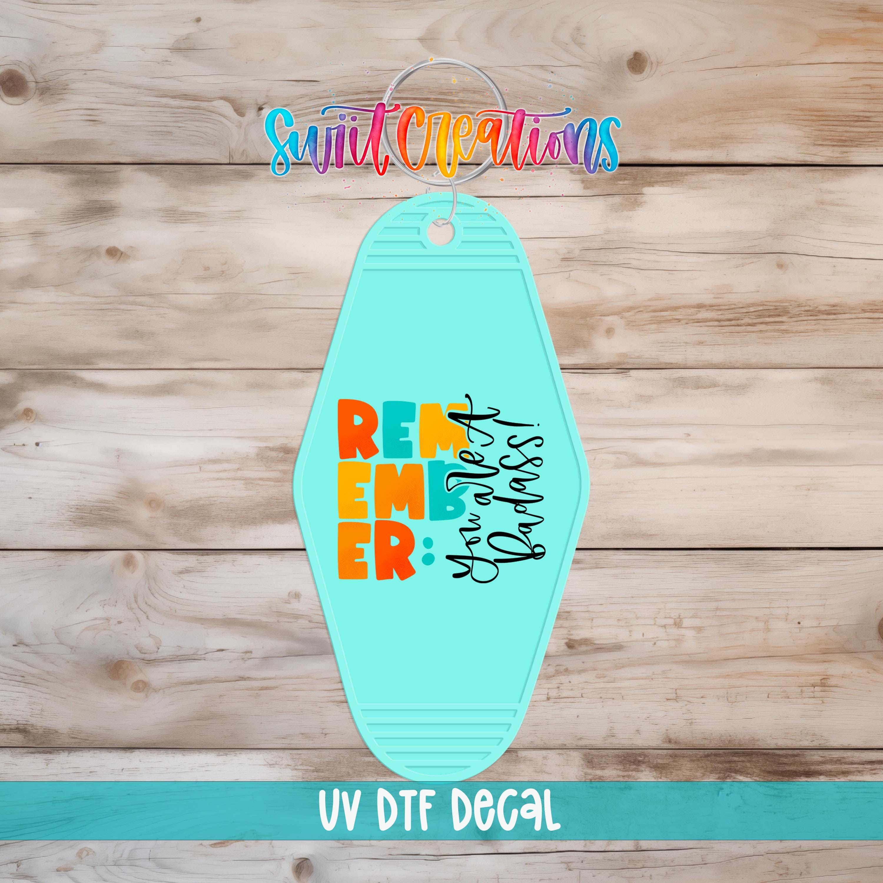 A turquoise keychain with the text "Remember" and "Badass" printed on it, placed on a wooden surface.