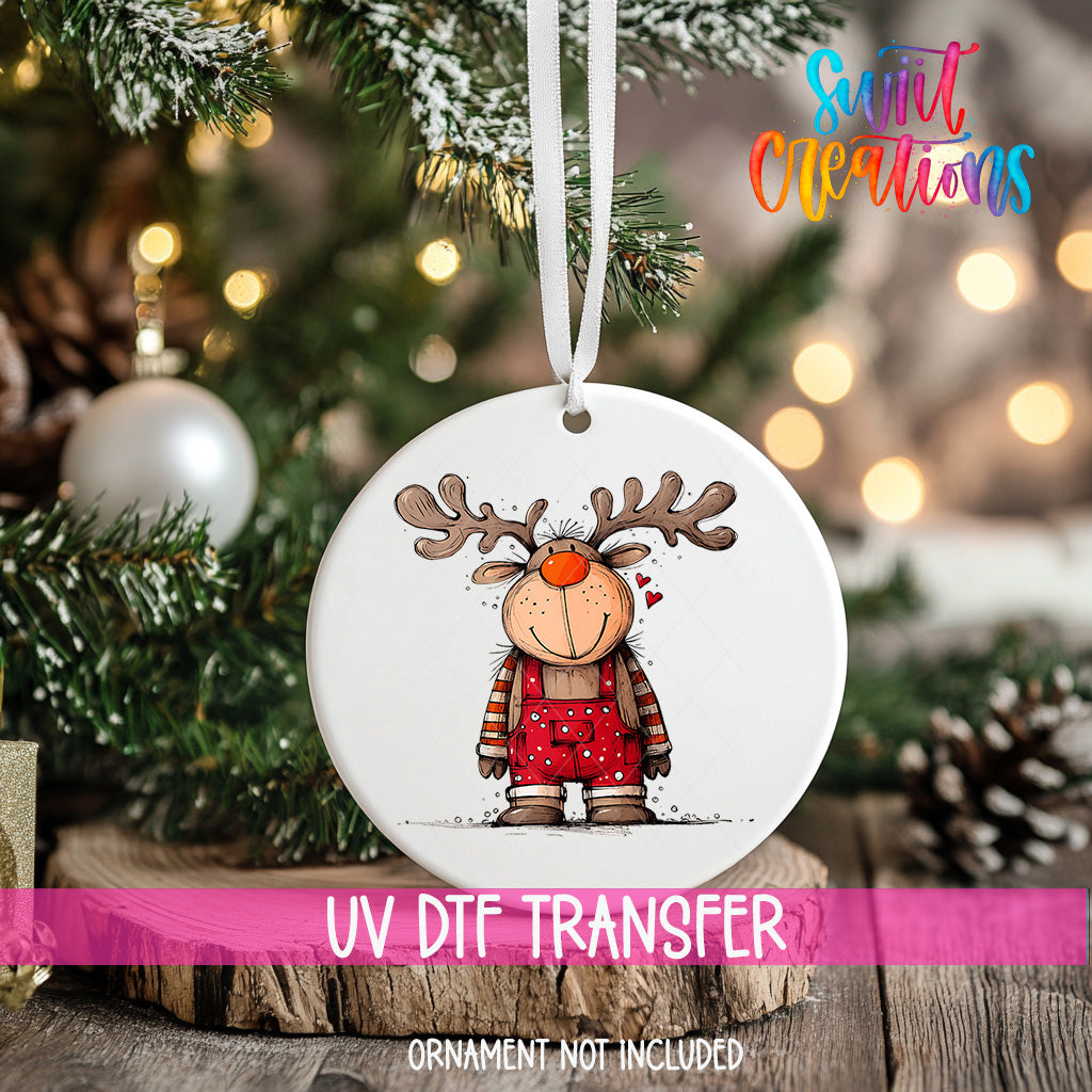 A white ornament with a cartoon reindeer design hangs on a Christmas tree, surrounded by pine branches and twinkling lights.