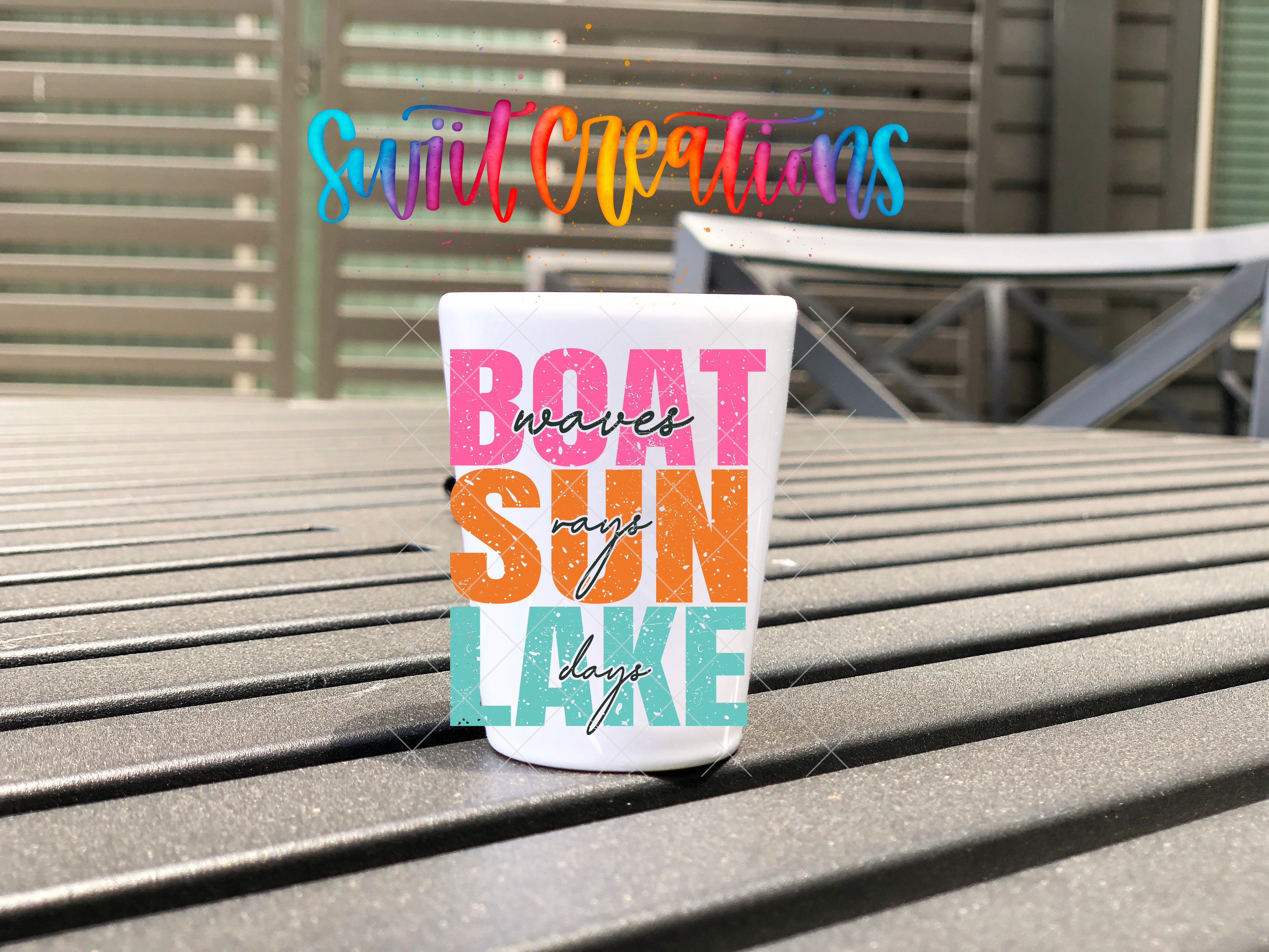 A colorful cup with text that says "BOAT SUN LAKE" is placed on a wooden surface, with a blurred background that includes a building and a railing.