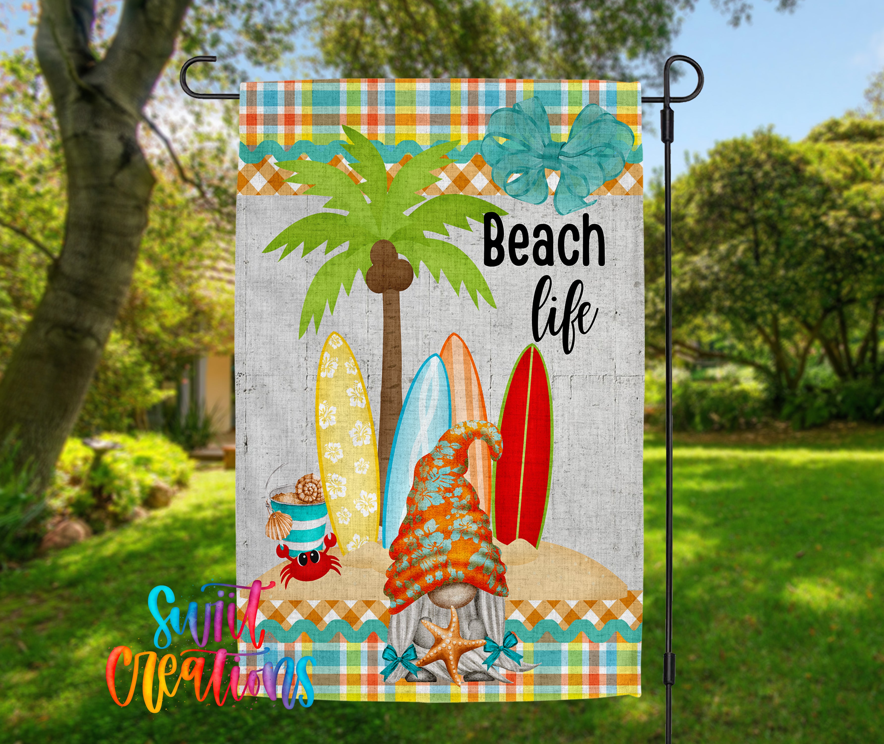 A colorful beach-themed garden flag with a gnome, palm tree, and surfboards, set against a backdrop of trees and a grassy lawn.
