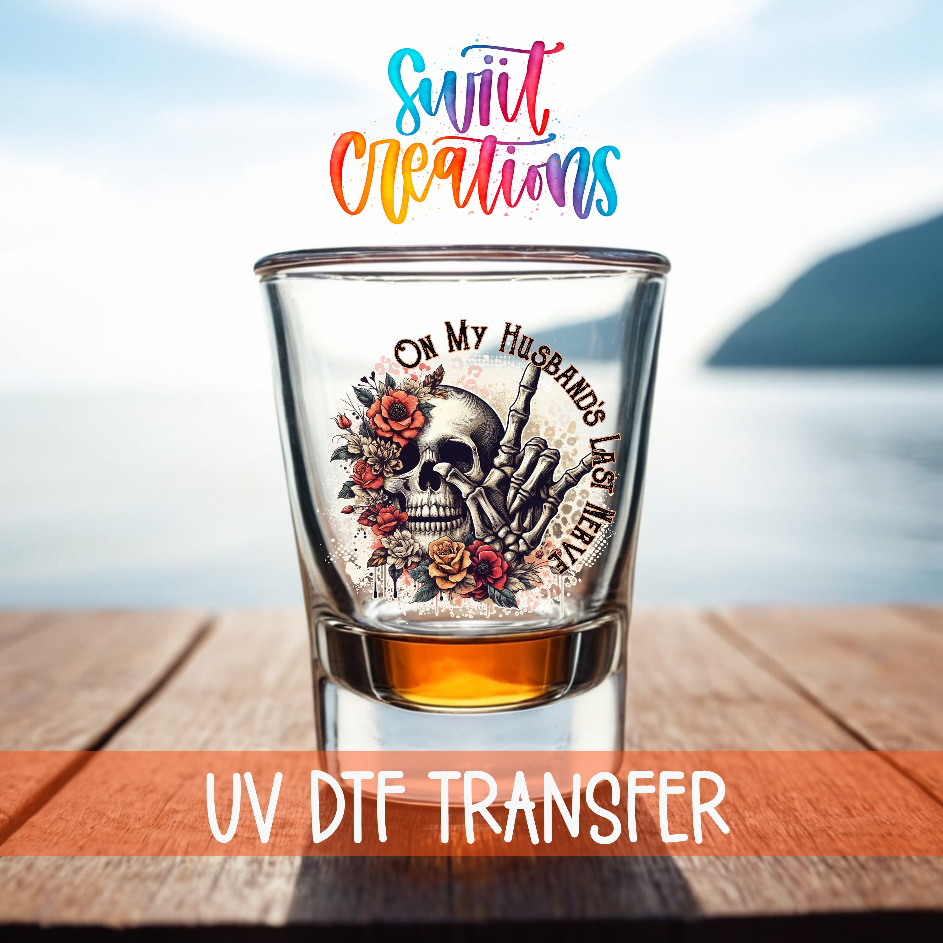 A shot glass with a skull design and floral elements, placed on a wooden surface with a body of water in the background.