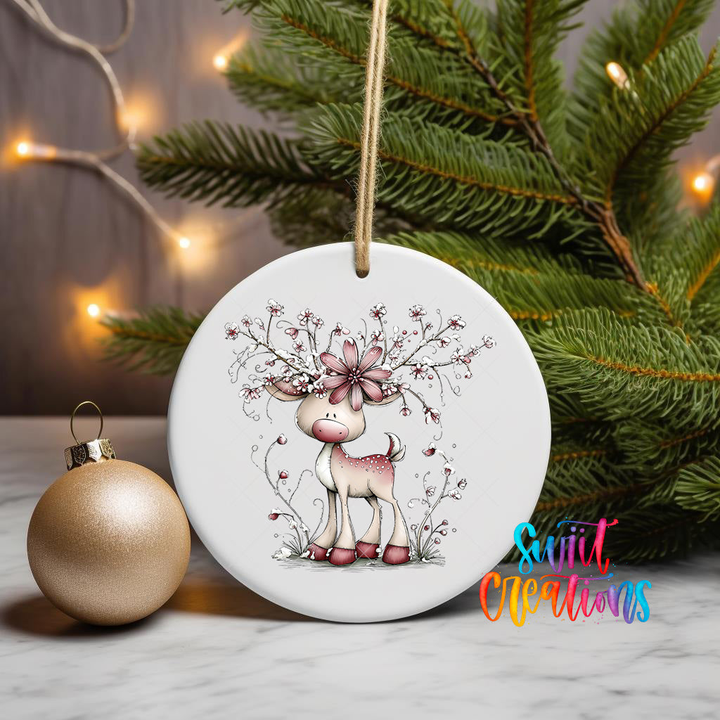 A white ceramic ornament featuring a deer with a flower on its head, surrounded by greenery, is displayed on a white surface next to a gold ornament.