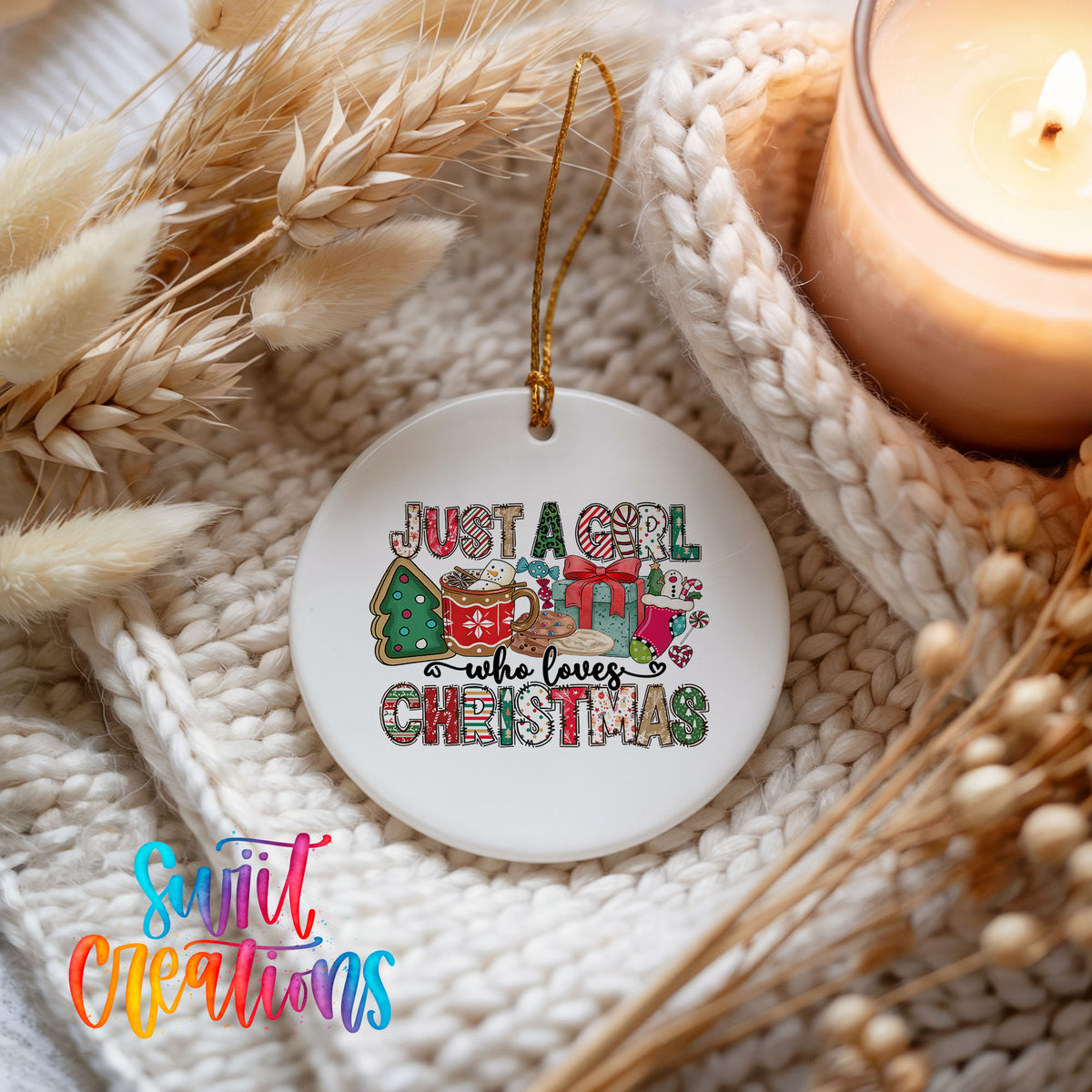 A white ceramic ornament with a colorful design hangs on a white knitted blanket, surrounded by dried wheat and a lit candle.