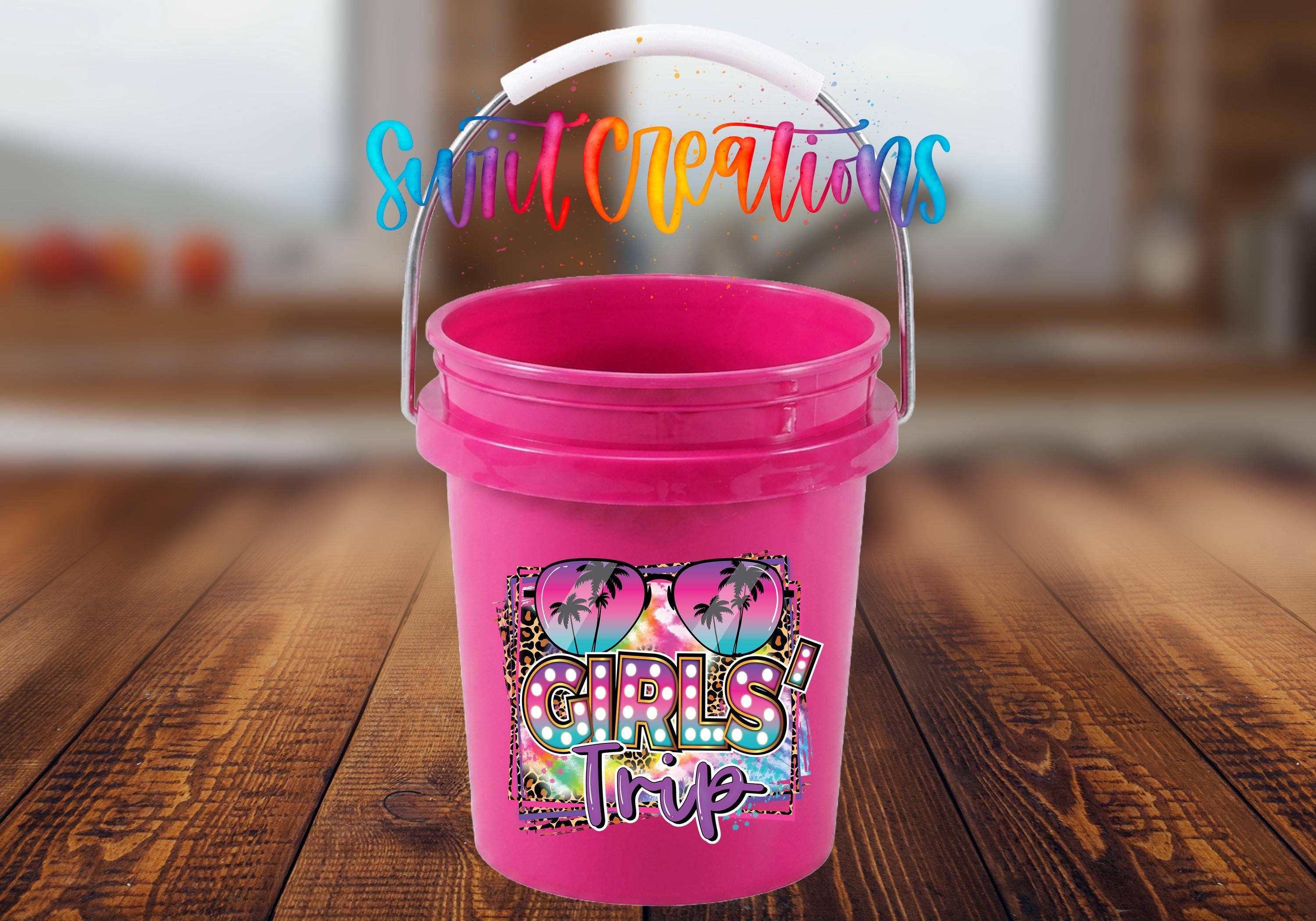 A pink bucket with a handle and a colorful design featuring the text "GIRLS TRIP" is displayed on a wooden surface.