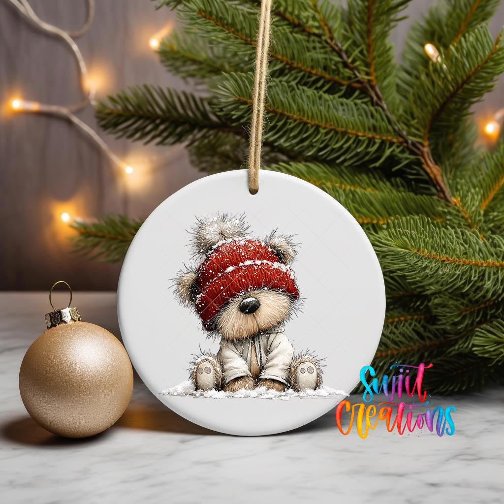 A white ceramic ornament featuring a teddy bear wearing a red hat and sitting in the snow, with a Christmas tree and twinkling lights in the background.