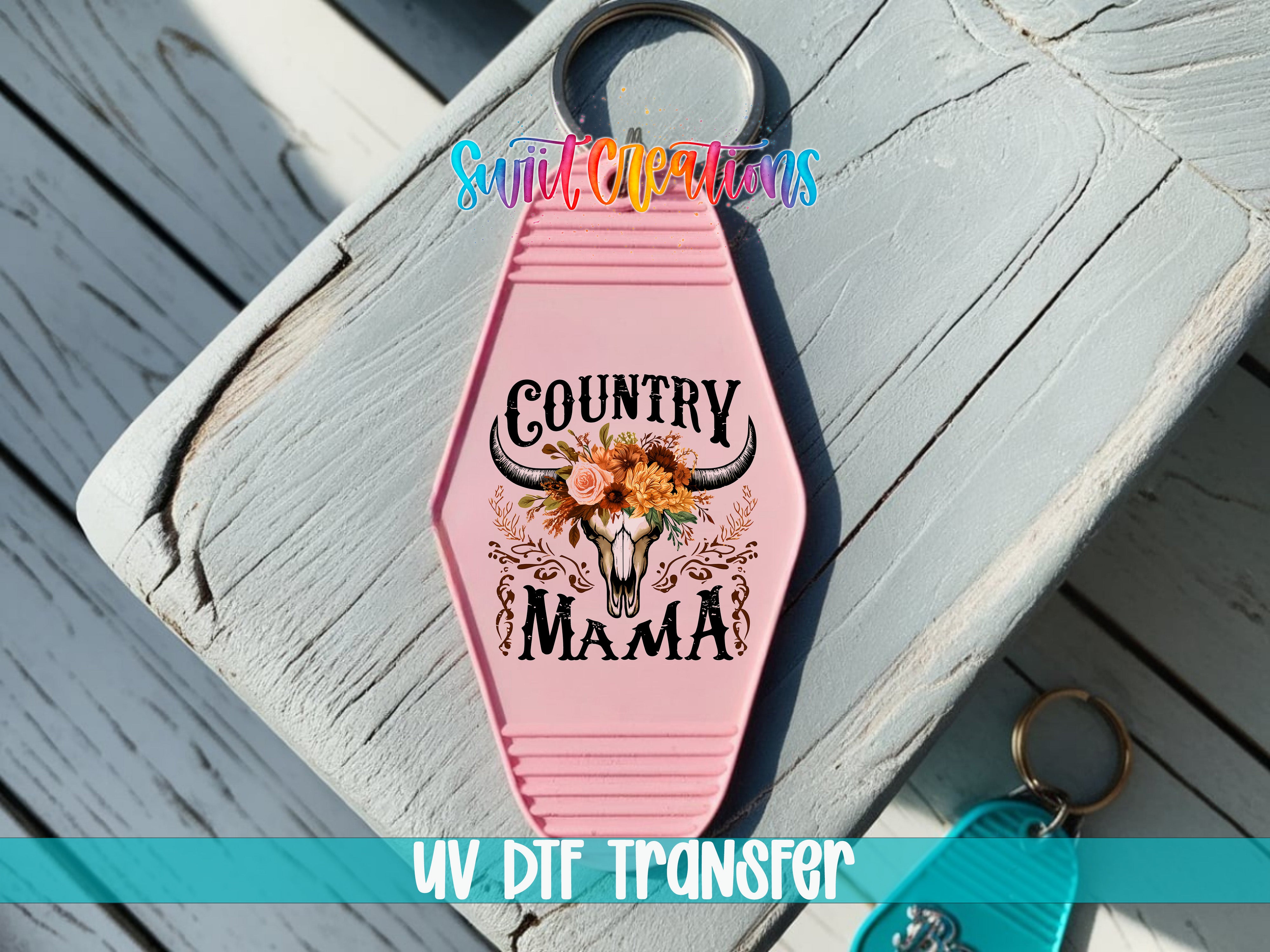 A pink keychain with a floral design and the text "COUNTRY MAMA" printed on it, placed on a wooden surface.