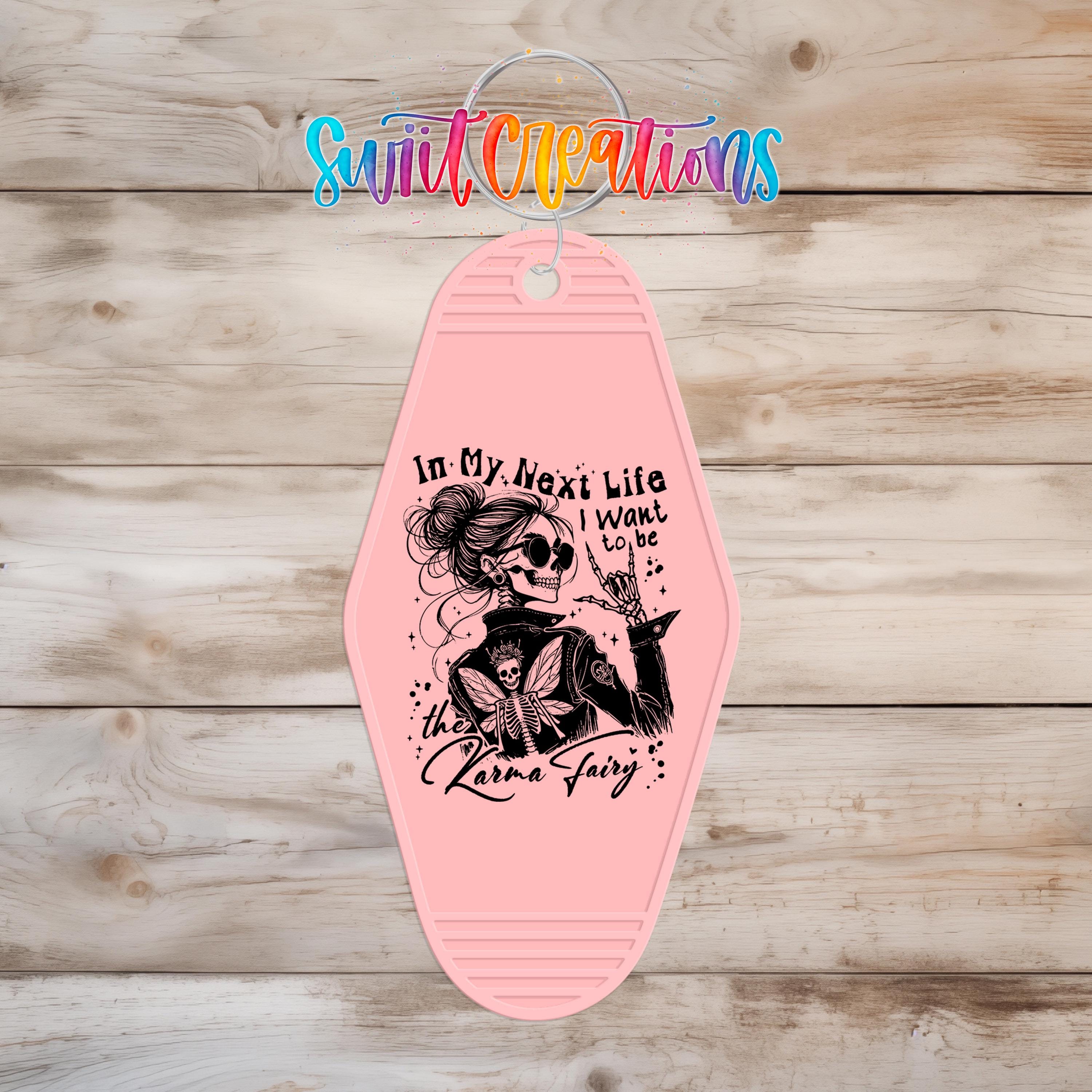 A pink keychain with a graphic design featuring a skeleton and a fairy-like figure, along with text that reads "In My Next Life I Want to Be the Karma Fairy".