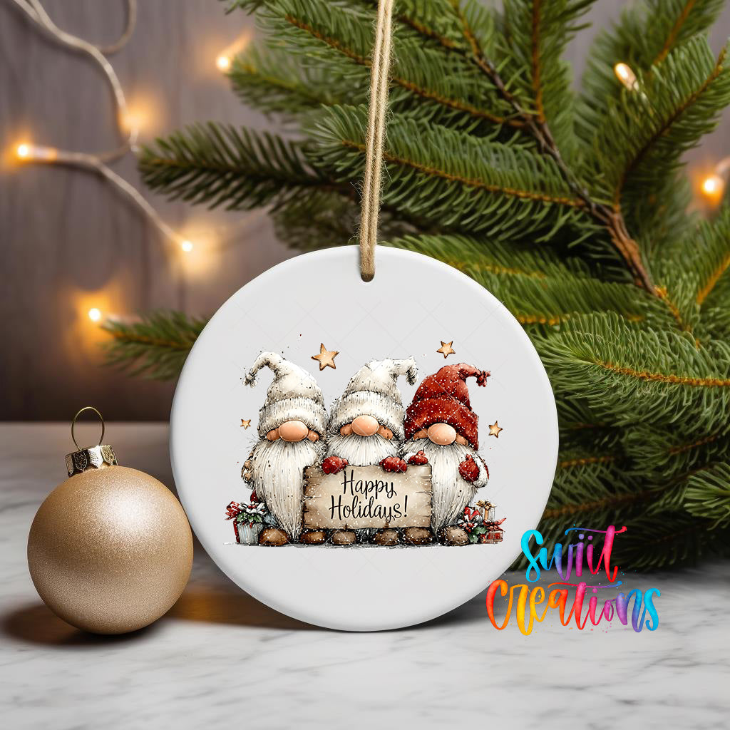 A white ceramic ornament with a festive design featuring three gnomes holding a sign that says "Happy Holidays!" is displayed on a table, with a Christmas tree and a gold ornament in the background.