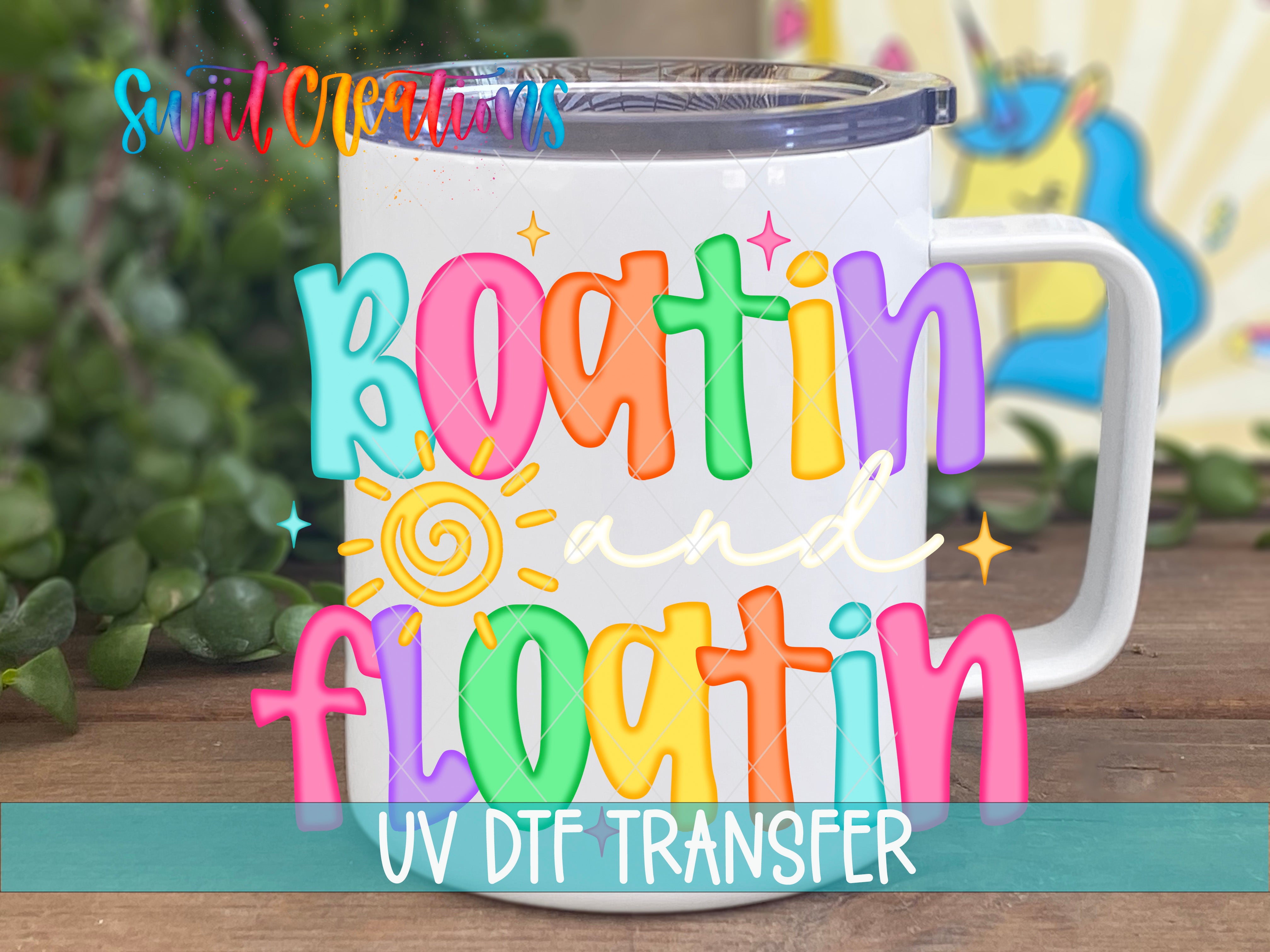 A colorful mug with the text "Boatin and floatin" printed on it, along with a sun and star design. The mug is placed on a wooden surface, and there is a blurred background that includes some greenery.