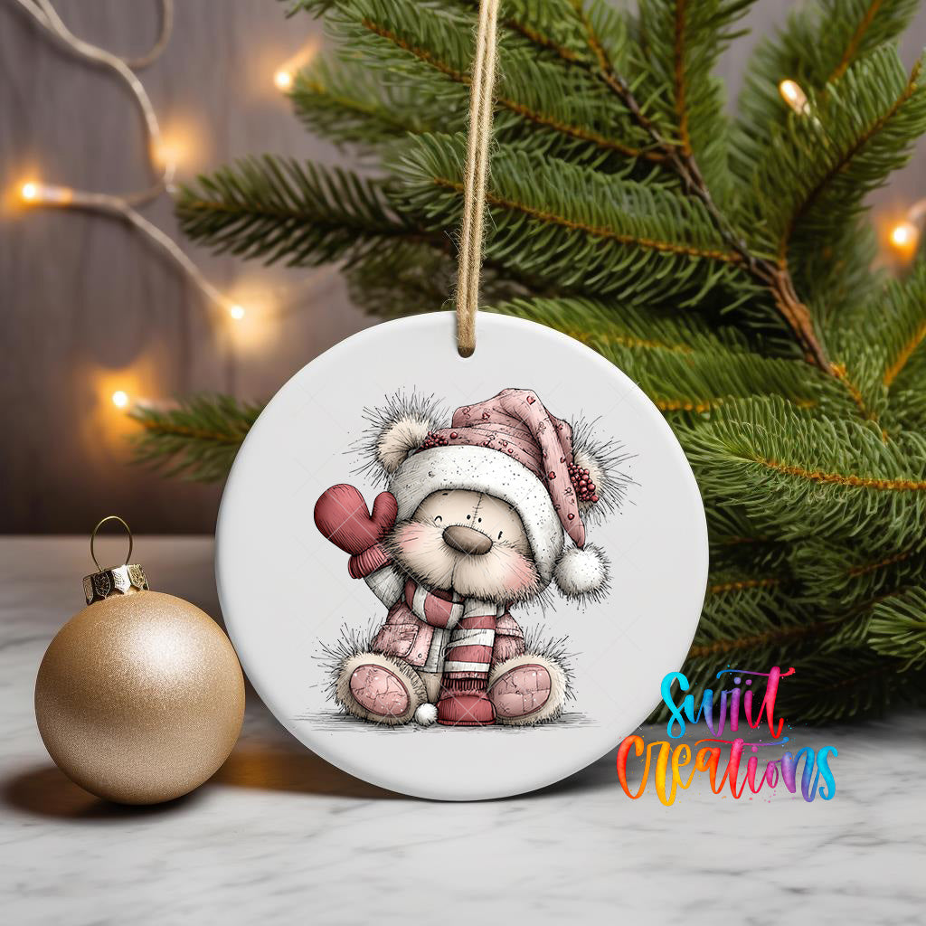 A white ceramic ornament featuring a teddy bear wearing a hat and scarf, sitting on a white surface next to a gold ornament and a Christmas tree with twinkling lights.