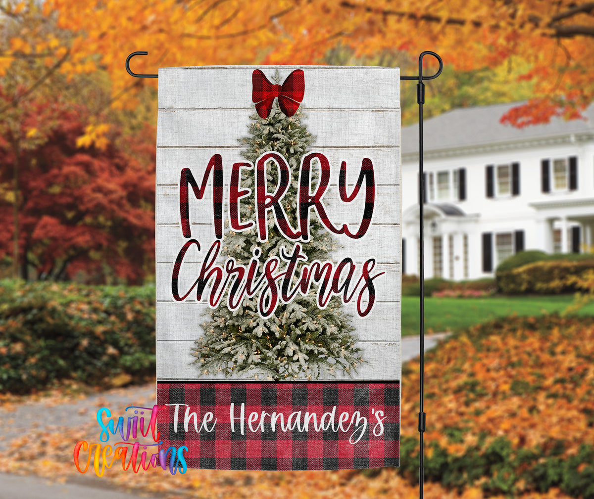 A white wooden Christmas banner with a red bow, displaying the text "MERRY CHRISTMAS" and "The Hernández's" in a colorful plaid pattern. The banner is attached to a black pole, with a house and autumn foliage visible in the background.