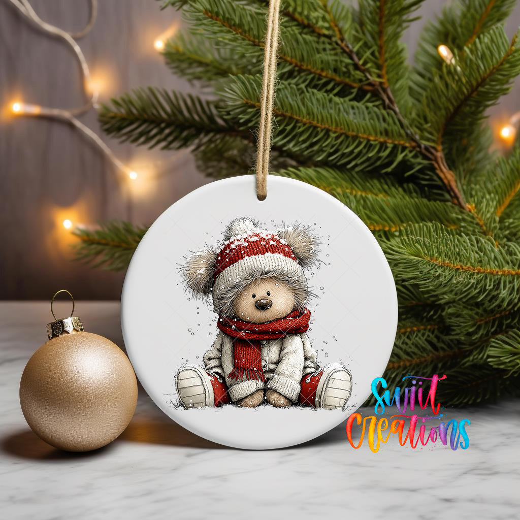 A white ceramic ornament featuring a teddy bear wearing a red scarf and hat, sitting on a white surface next to a gold ornament and a Christmas tree.