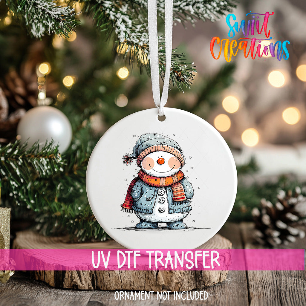 A white ornament with a snowman design hangs on a Christmas tree, surrounded by pine branches and twinkling lights.