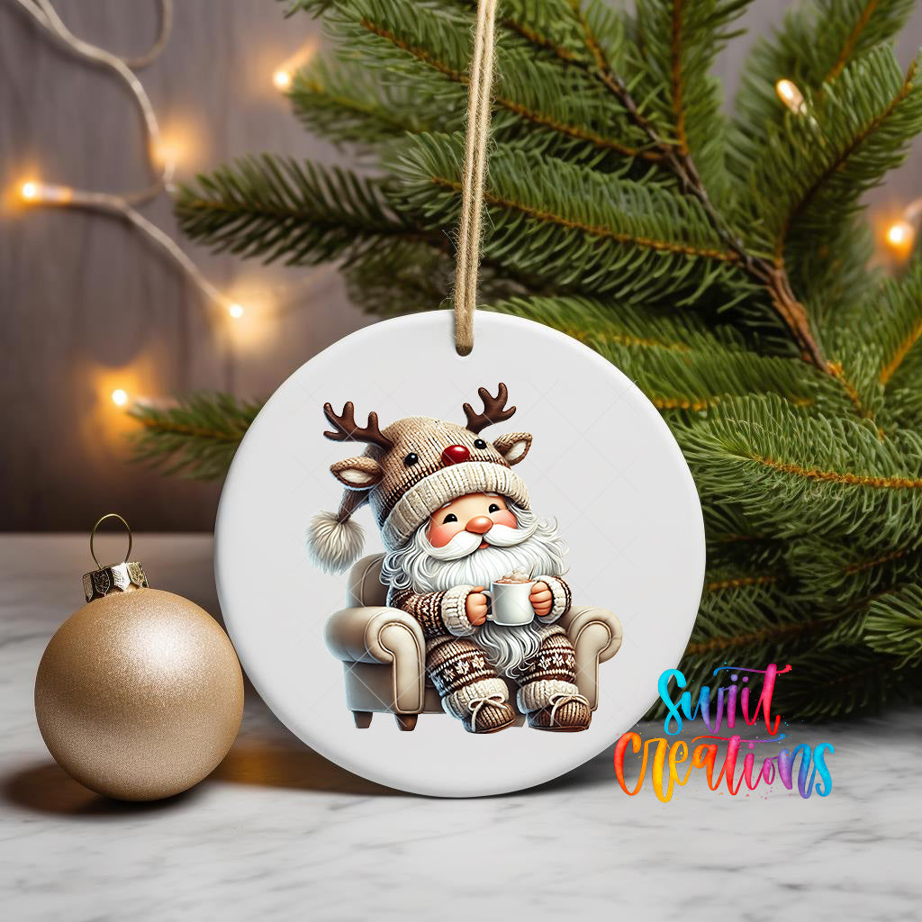 A white ornament featuring a cartoon Santa Claus sitting in a chair, wearing a reindeer hat and holding a cup, is displayed on a table next to a gold ornament and a Christmas tree.