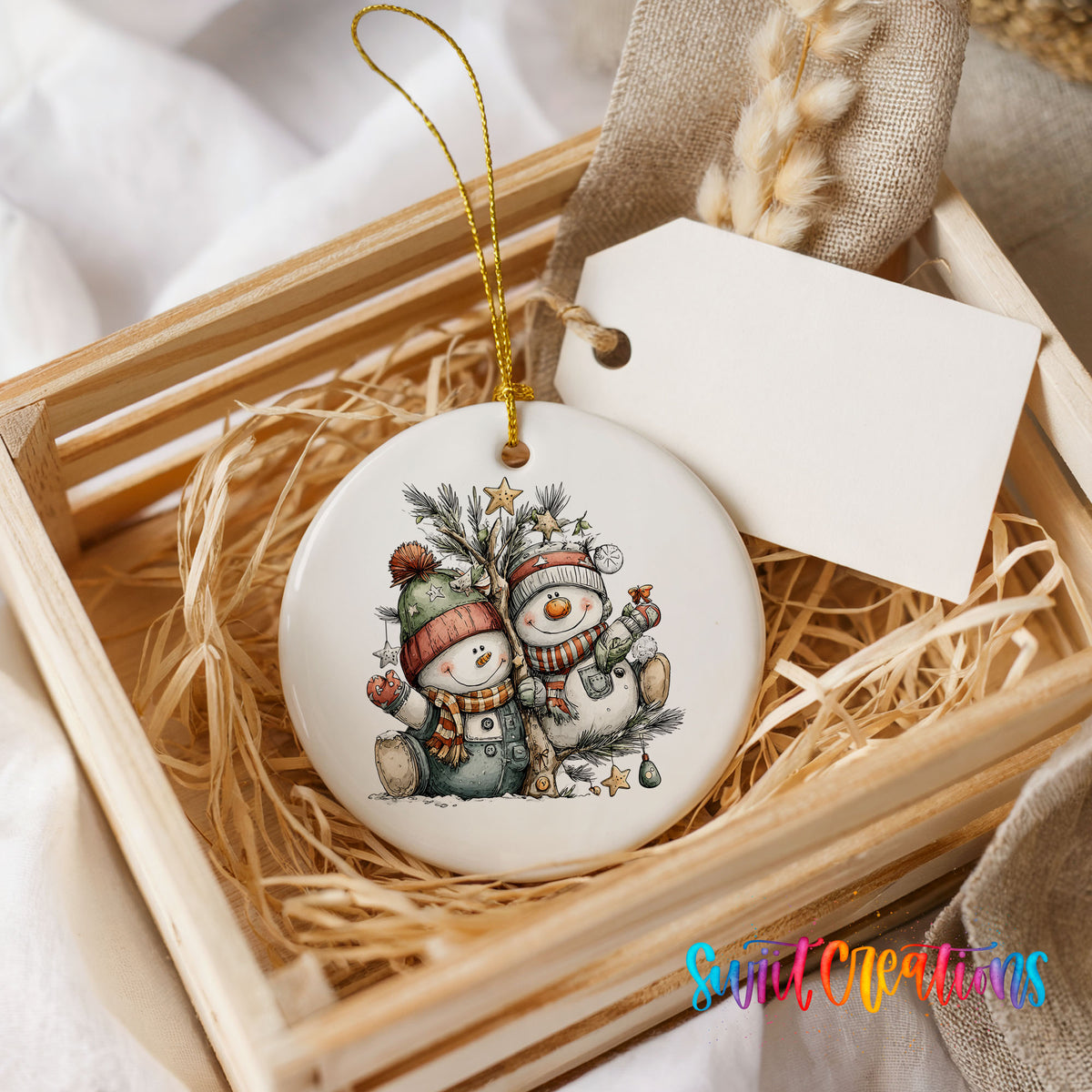 A ceramic ornament featuring two snowmen is displayed in a wooden crate, with a white tag attached to it.
