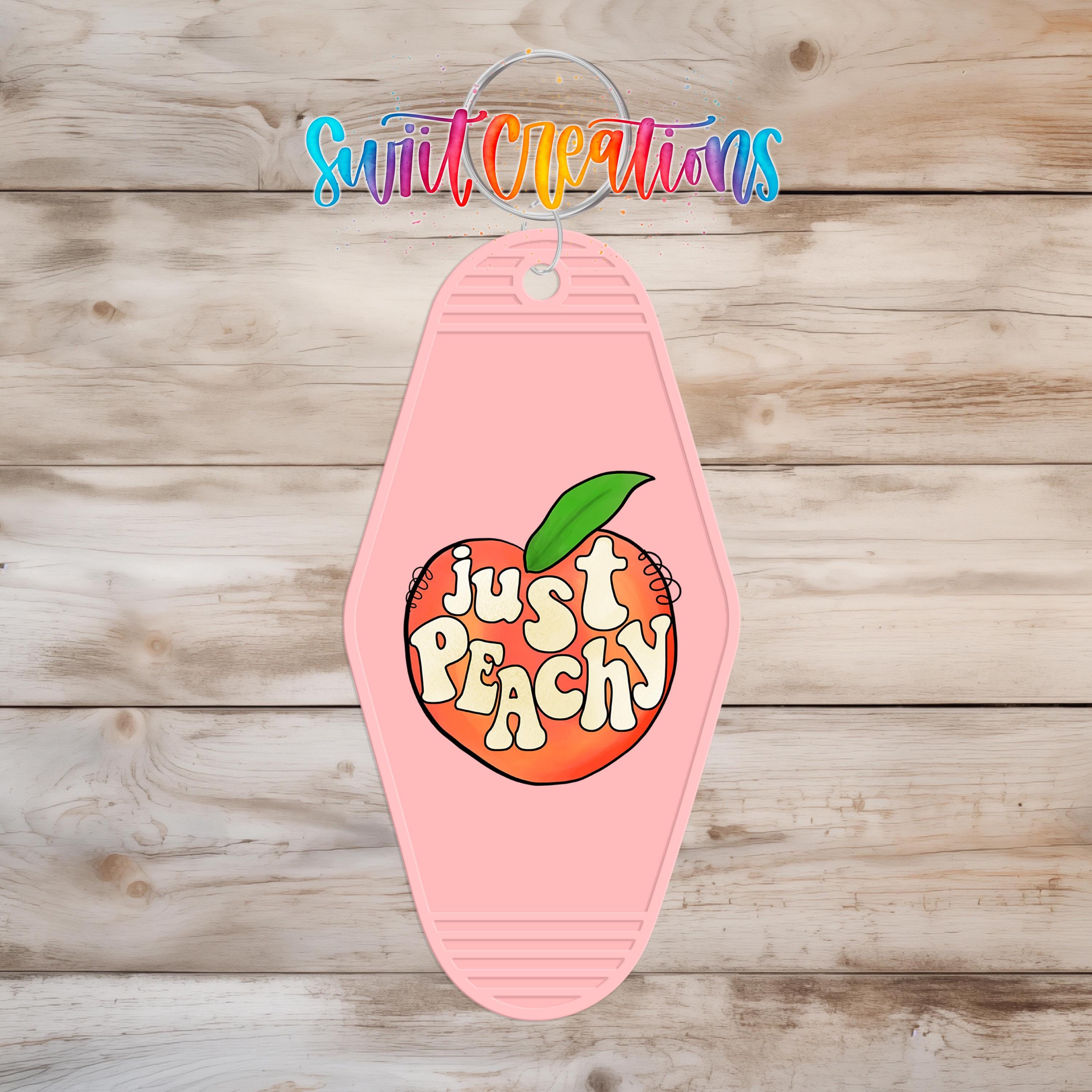 A pink keychain with a peach graphic and the text "Just Peachy" printed on it, hanging on a wooden surface.