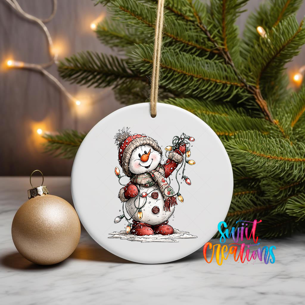 A white ornament featuring a snowman is displayed on a table, with a Christmas tree and a gold ornament visible in the background.