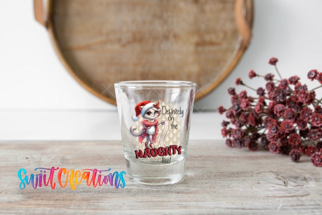 A shot glass with a cartoon cat wearing a Santa hat and the text "Despondency of the Naughty" printed on it, placed on a wooden surface next to a bunch of red flowers.
