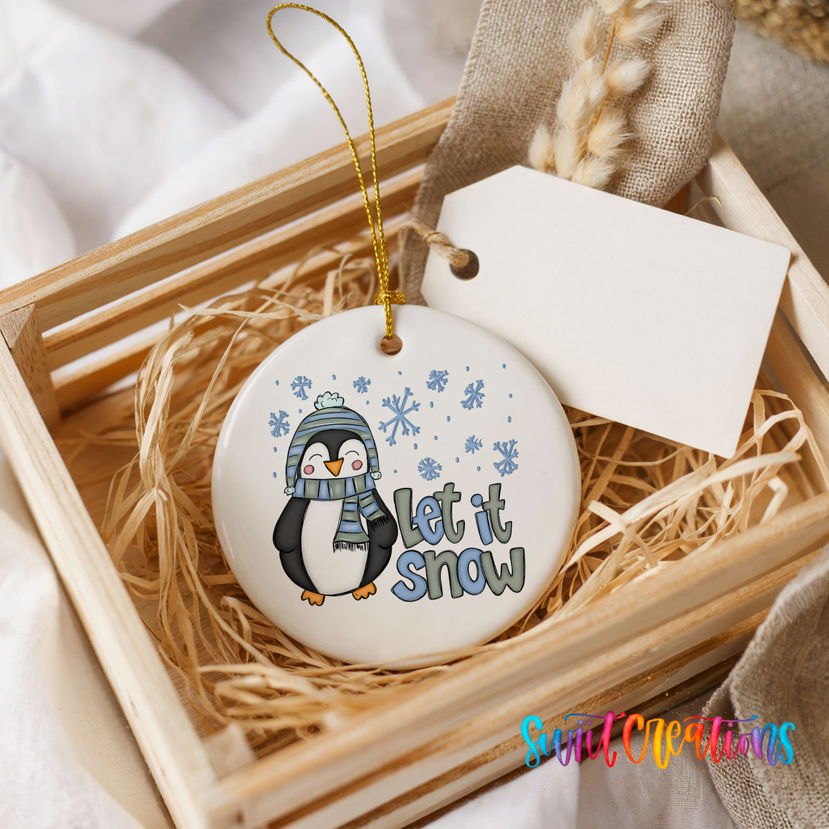 A white ceramic ornament with a penguin design and the text "Let it snow" is displayed in a wooden crate.