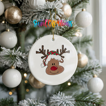 A white ceramic ornament with a cartoon reindeer wearing a Santa hat is hanging on a Christmas tree.