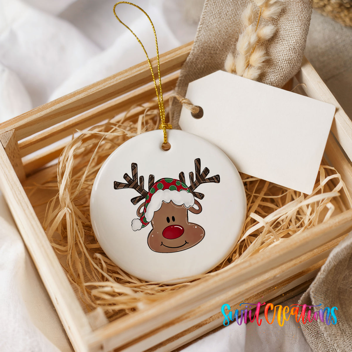 A white ceramic ornament with a reindeer face design is displayed in a wooden crate, accompanied by a white tag and straw.