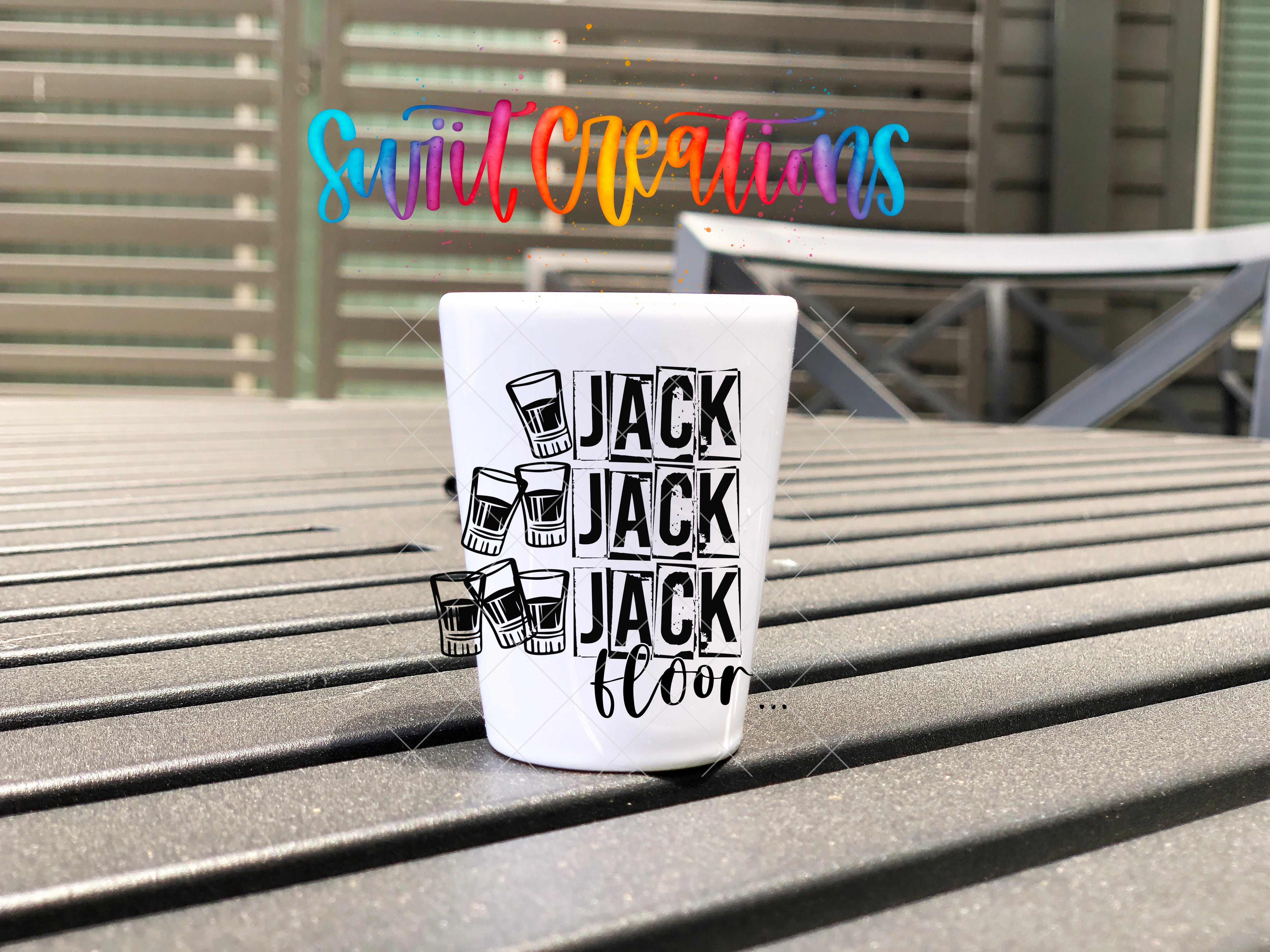 A white shot glass with the text "JACK JACK JACK JACK" printed on it, placed on a wooden surface.