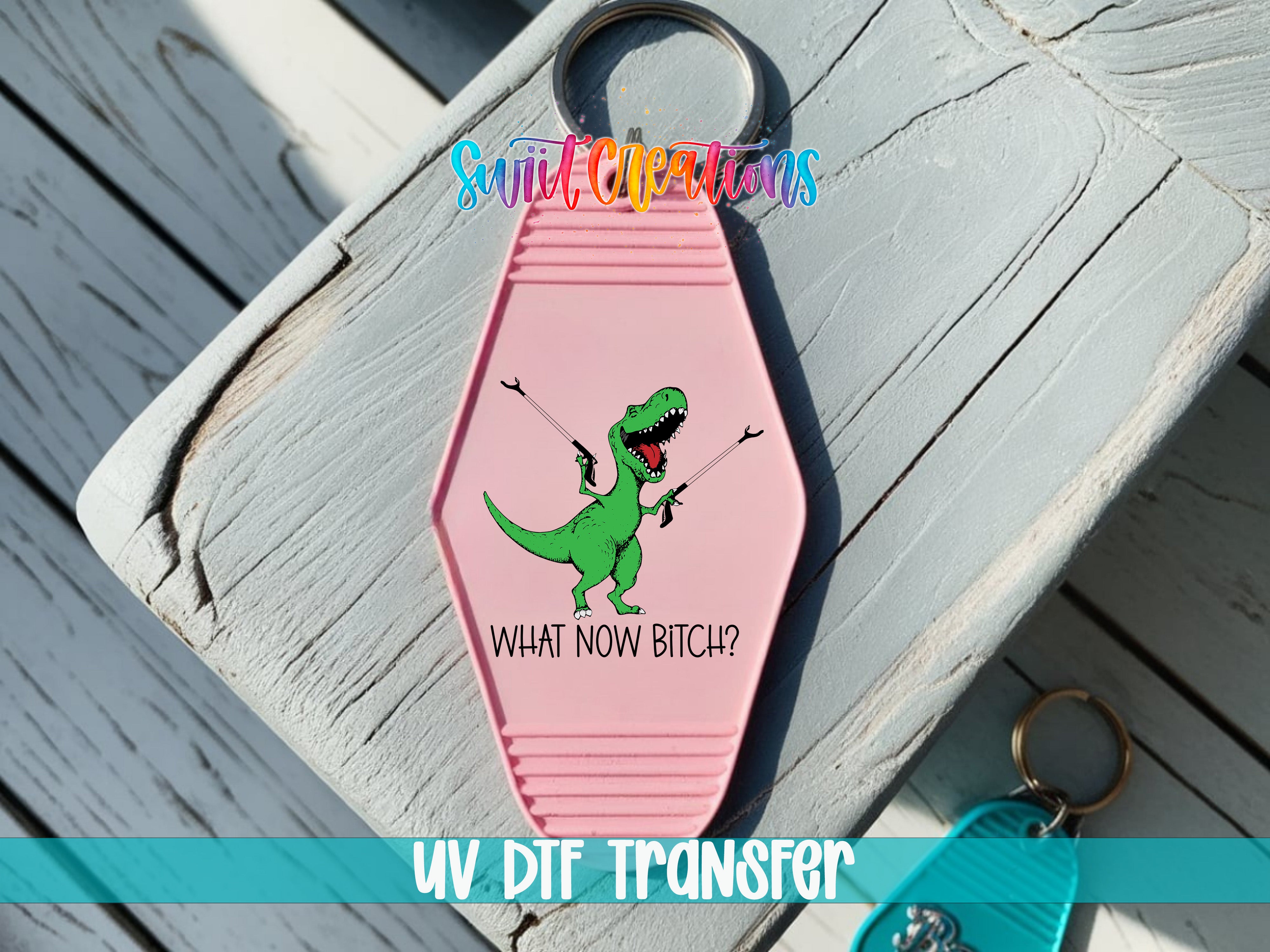 A pink keychain with a cartoon dinosaur and the text "WHAT NOW BITCH?" printed on it, placed on a wooden surface.