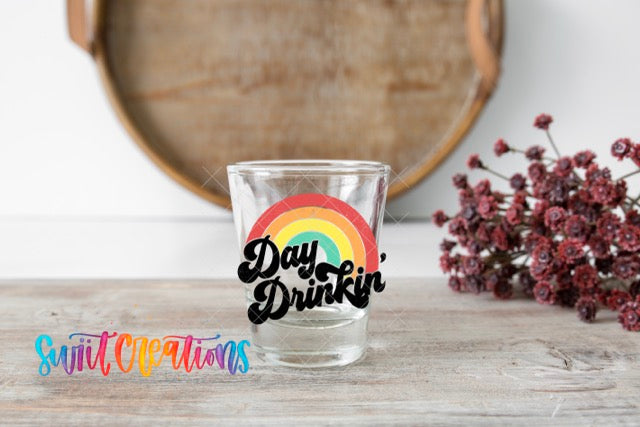 A glass with a rainbow-colored design and the text "Day Drinkin'" sits on a wooden surface, with a bunch of red flowers in the background.